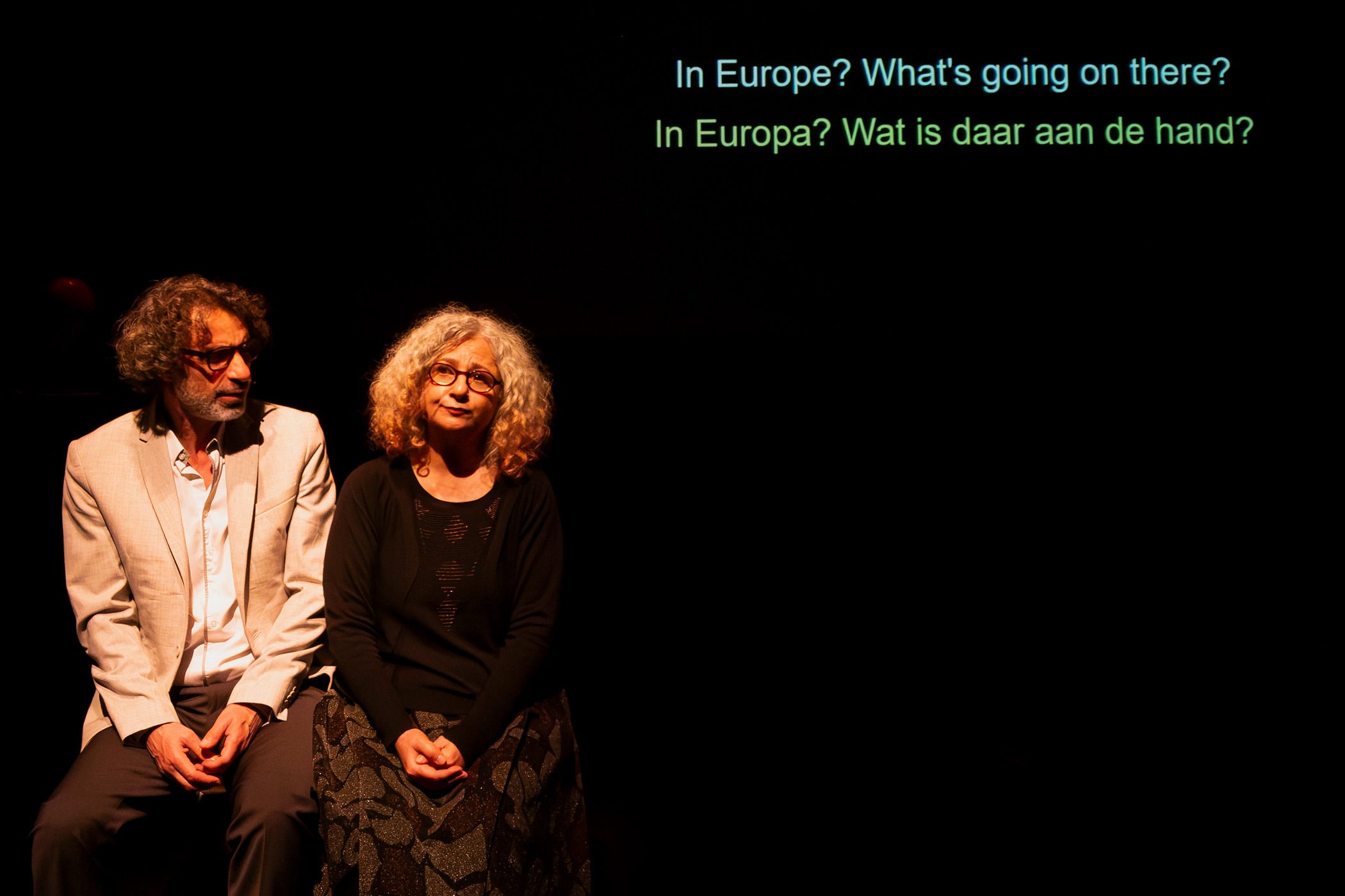 Majdalanie-Mroue_Four-Walls-and-a-Roof_c_Sybille-Cornet_02WEB Zwei Personen sitzen auf einer Bank und sprechen miteinander. An der Wand im Hintergrund ist Schrift Projiziert.